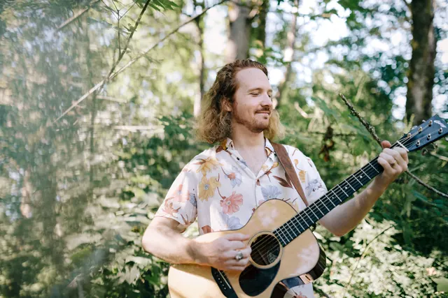 Ben playing acoustic guitar among the trees.