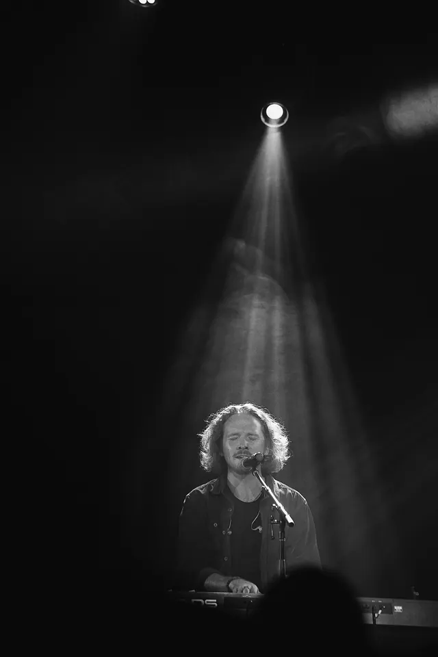 Ben singing and playing keys beneath a spotlight in a black and white stage photograph.